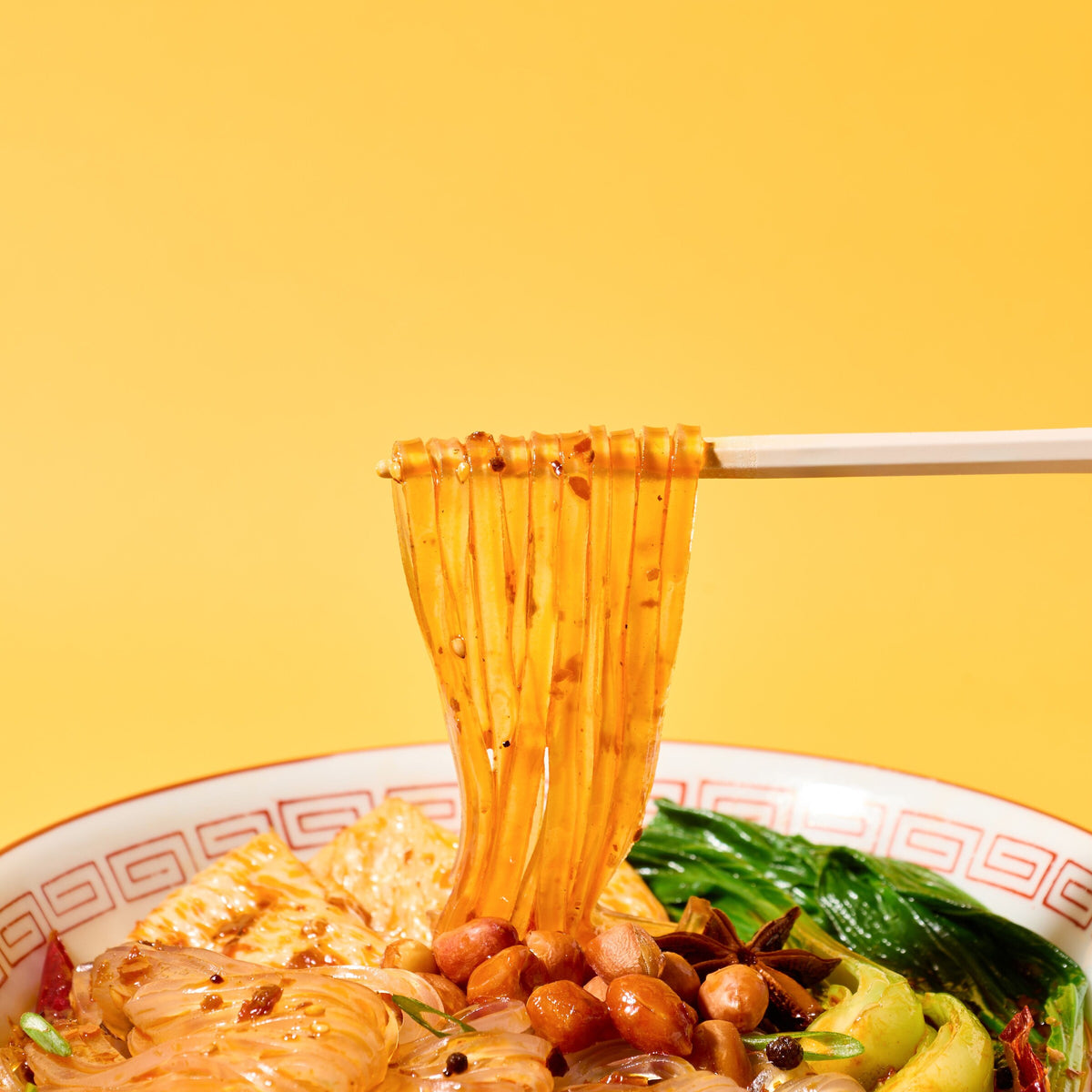 Chopsticks lifting glossy kuzukiri glass noodles from a spicy bowl